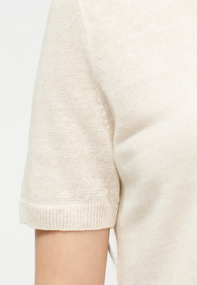 Gros plan sur le haut du bras d'une personne portant un pull en maille beige clair à manches courtes avec un poignet côtelé.