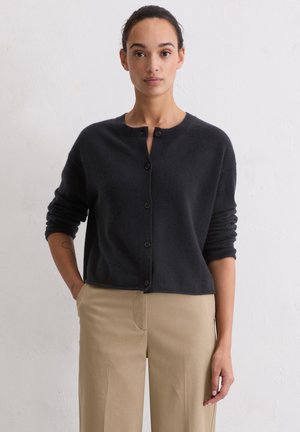 Donkergrijze cardigan van zacht materiaal, met een ronde halslijn, vijf knopen aan de voorkant en opgerolde mouwen. Gecombineerd met beige pantalon.