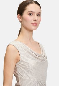 Sukienka z dekoltem w serek w jasnym metalicznym materiale o teksturowanym wykończeniu. Bezrękawnik z delikatnym połyskiem i drapowaniem przy dekolcie.