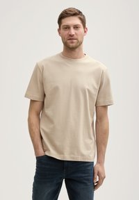 Beige, rövid ujjú póló, puha pamut anyagból, kerek nyakkivágással és laza szabással, sötétkék farmernadrággal viselve.