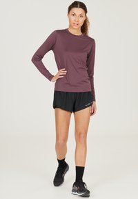 Langärmliges, kastanienbraunes Sportoberteil, kombiniert mit schwarzen Shorts. Das Oberteil hat einen runden Halsausschnitt, und die Shorts verfügen über einen elastischen Bund und ein kleines Logo.