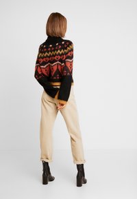 Pull en tricot avec des motifs géométriques rouges, noirs et jaunes, associé à un pantalon beige clair et des bottes à talons noires, vu de dos.