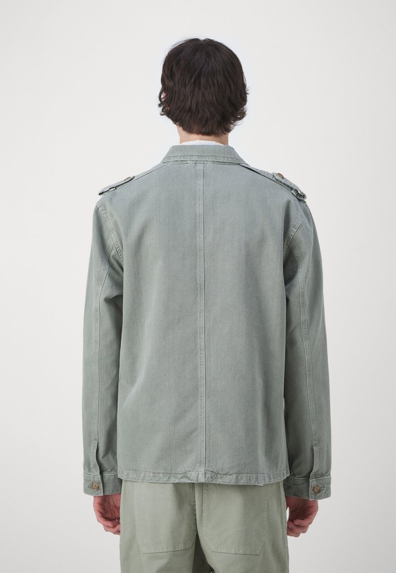 Chaqueta de mezclilla verde clara con corte recto, epaulettes en los hombros y acentos de botones. Tela texturizada con mangas largas y un detalle de costura en la espalda.