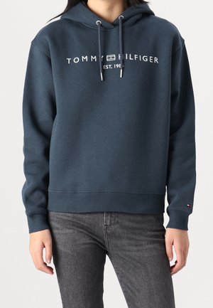 Person trägt einen dunkelblauen Tommy Hilfiger Hoodie mit weißen Kordeln und Schriftzug, kombiniert mit dunkelgrauen Jeans.