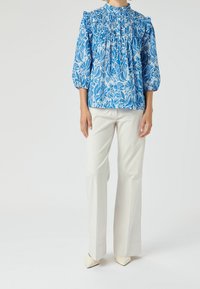 Blusa floreale blu con spalle arricciate, scollatura arricciata e maniche a palloncino, abbinata a pantaloni bianchi a gamba dritta e tacchi color crema.