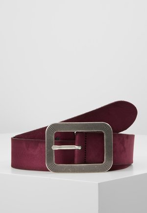 Laaja tummanpunainen mokkanahkavyö, jossa on suuri suorakaiteen muotoinen hopeinen teksturoitu solki, kierrettynä valkoisen jalustan päällä vaaleaa taustaa vasten.