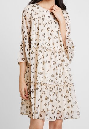 Kobieta w luźnej, beżowej sukience sięgającej do kolan, z warstwowymi falbanami i metalicznym brązowym wzorem w cętki, z ręką opartą blisko szyi.