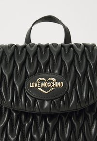 Schwarze, gestepptes Lederhandtasche mit einem strukturierten, wellenförmigen Design, ausgestattet mit einer goldfarbenen Logo-Plakette und einem oberen Tragegriff.