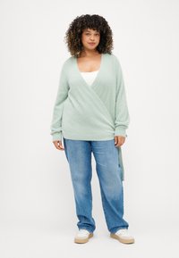 Ανοιχτό πράσινο περιτύλιγμα κάρντιγκ από υφή υφάσματος, με μακριά μανίκια, βαθύ V λαιμόκοψη και μακρύτερο πίσω στρίφωμα. Συνδυάζεται με ανοιχτό γαλάζιο τζιν.