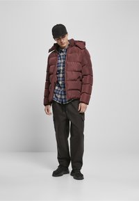 Chaqueta de plumas granate con capucha, que presenta una cremallera y costuras horizontales, llevada sobre una camisa a cuadros azul, combinada con pantalones cargo oscuros y zapatillas negras.