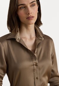 Brūnas satīna blūze ar pogu aizdari un lielu, uzpūstu apkakli. Gluda tekstūra un pieguļošs dizains uzlabo tās eleganto izskatu.