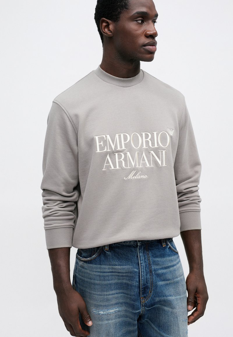 Sweatshirt gris clair avec col rond, arborant un logo brodé en blanc "EMPORIO ARMANI Milano". Porté avec un jean bleu déchiré.