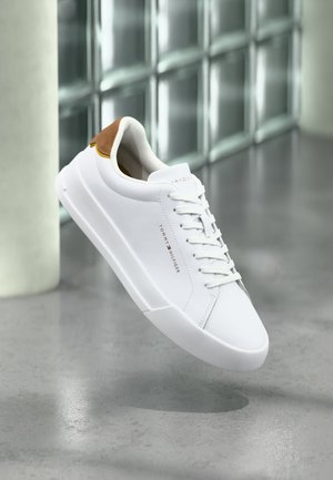 Άσπρο χαμηλό παπούτσι με ταμπ για τη φτέρνα από καφέ σουέτ, άσπρα κορδόνια και λογότυπο Tommy Hilfiger στο πλάι, να αιωρείται πάνω από ένα γυαλισμένο γκρι πάτωμα.