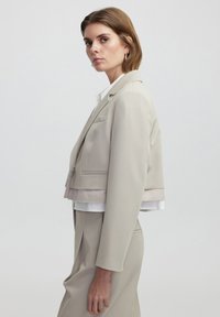 Giacca beige tagliata con una texture liscia, rever affilati e una tasca singola, sovrapposta a una maglietta bianca con dettagli visibili sull'orlo.