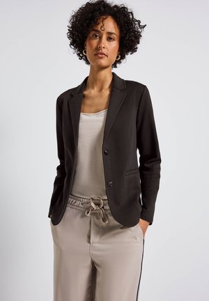 Zwarte blazer met puntige revers, sluiting met één knoop en twee voorkwakken, gecombineerd met een lichtbeige tanktop en bijpassende broek.