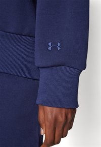 Marineblå sweaterstof med ribbet manschette, prydet med et mørkeblåt broderet Under Armour-logo på håndledsområdet.