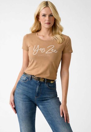 Donna bionda che indossa una maglietta color tan "Yes Zee Natural Feeling", jeans blu e cintura nera con fibbia dorata, in piedi su sfondo bianco.