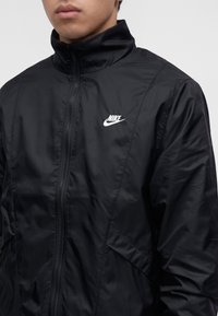 Νέος άνθρωπος που φοράει μαύρο μπουφάν Nike με φερμουάρ, ψηλό γιακά και λευκό λογότυπο Nike στο στήθος, στραμμένος ελαφρώς προς τα αριστερά.