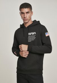 Un sweat à capuche noir avec le logo de la NASA et du texte de mission imprimé en blanc. Il possède une poche avant et un patch du drapeau américain sur la manche gauche.