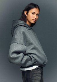 Sivá mikina s kapucňou s textúrovaným, lesklým povrchom. Vyznačuje sa nadrozmerným strihom a rebrovanými manžetami, doplnená tmavými rifľami.