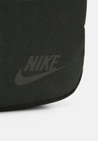Grön tygväska med en strukturerad yta som har en präglad svart Nike-logotyp och swooshdetalj på framsidan.