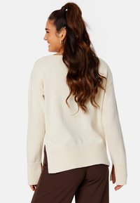 Maglione lavorato a maglia di colore crema con spacchi laterali e polsini a coste, caratterizzato da una vestibilità morbida e finitura testurizzata. Abbinato a pantaloni marroni.