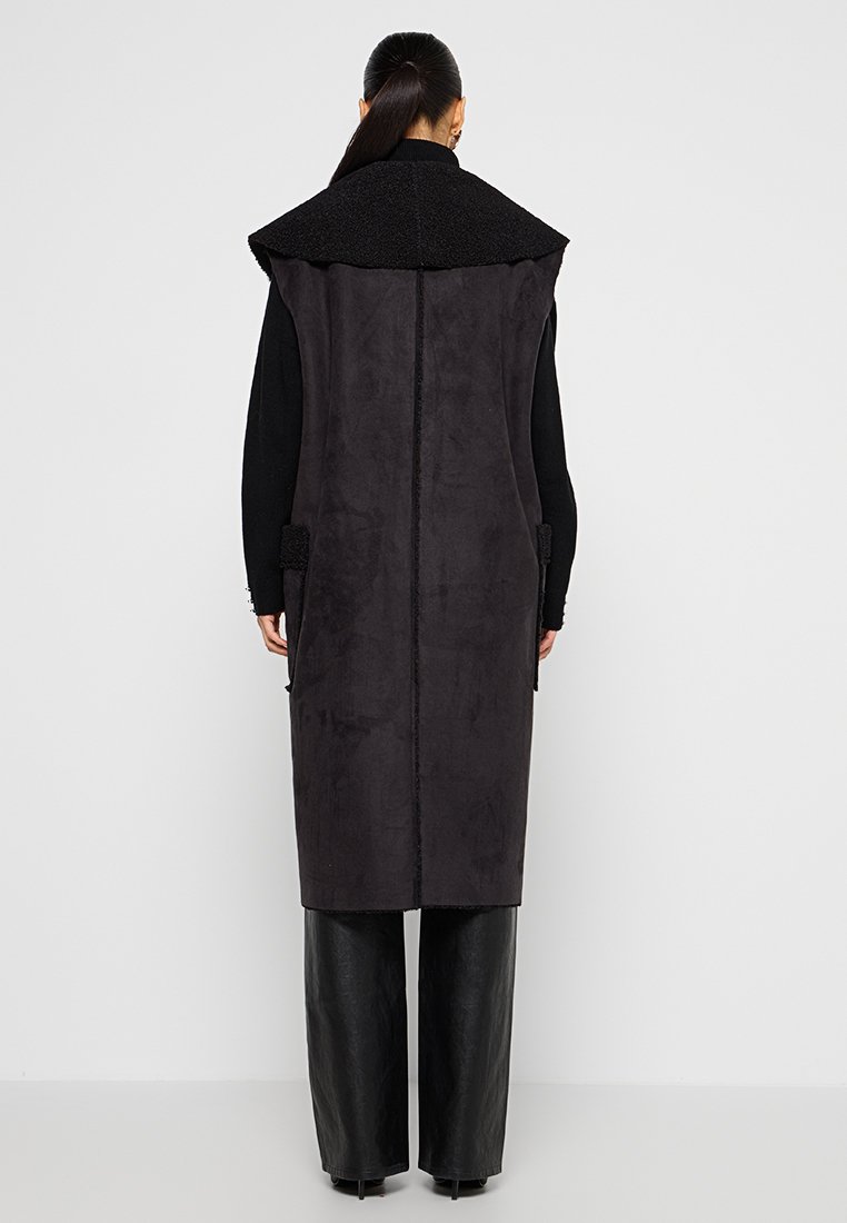 Langer schwarzer ärmelloser Mantel mit texturiertem Schalkragen, seitlichen Taschen und glatter Oberfläche, kombiniert mit weiten schwarzen Hosen.