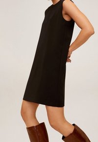 Robe noire sans manches, longueur genoux, portée avec des bottes en cuir marron jusqu'aux genoux, présentée sur un modèle contre un fond uni.