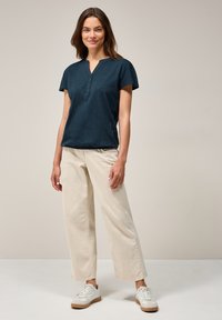 Camicetta navy a maniche corte con scollo a V e dettaglio dei bottoni, abbinata a pantaloni beige larghi e sneakers bianche, su uno sfondo neutro.