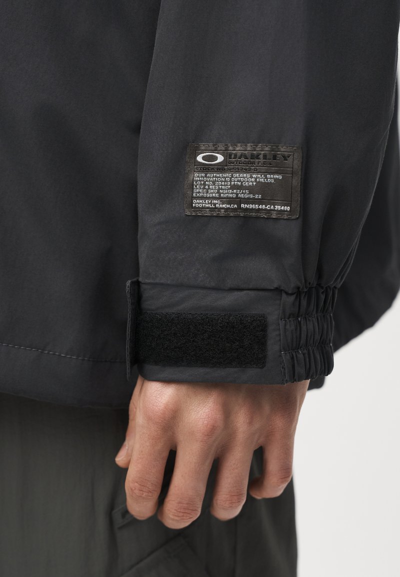 Primer plano de una mano que lleva una chaqueta negra con una etiqueta de Oakley y una correa de velcro en el puño de la manga.