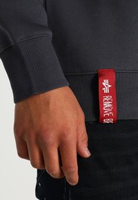 Una mano maschile appoggiata vicino al polsino di una felpa grigia scura con un'etichetta rossa che recita "RIMUOVI PRIMA DEL VOLO."