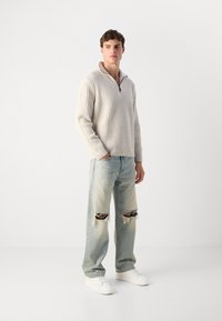 Hellbeige gerippter Pullover mit Reißverschlusskragen, kombiniert mit hellblauen, ausgewaschenen Jeans mit großen Rissen an den Knien und weißen Schuhen.