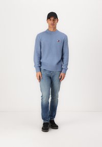 Blauwe geribde trui met ronde hals, gedragen met lichtblauwe jeans. Een zwarte pet en sneakers maken de outfit compleet. Klein logodetail op de trui.