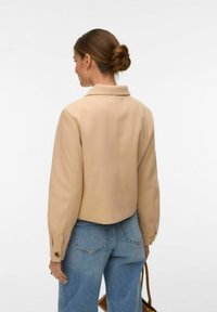 Femme avec un chignon bas, portant une veste beige à manches longues et un jean bleu, tenant un sac marron, vue de dos.