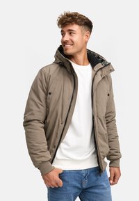 Ung mand iført en beige hættejakke over en hvid sweatshirt og blå jeans, som ser til siden med et smil.