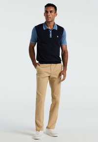 Marine blauwe vest over een lichtblauw overhemd met kraag, gecombineerd met khaki broek en witte sneakers. Bevat een klein logo op het vest.