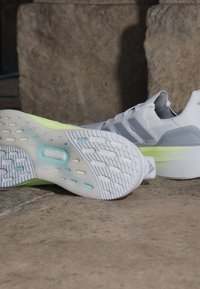 Paire de chaussures de sport blanches avec des semelles intermédiaires vert clair placées sur une surface en pierre, l'une montrant la semelle texturée et l'autre debout.