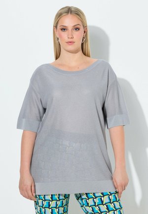 Junge Frau mit glattem blondem Haar trägt einen weiten hellgrauen Pullover mit kurzen Ärmeln und bunte gemusterte Hosen, steht vor einer weißen Wand.