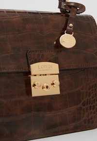 Sac en cuir à motif crocodile marron foncé avec une serrure rectangulaire dorée et une étiquette de marque ronde dorée attachée à la sangle.