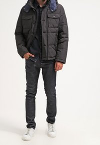 Ciemna kurtka puchowa z niebieskim kołnierzem, wyposażona w kilka przednich kieszeni, noszona na czarnej koszuli, zestawiona z ciemnymi dżinsami i białymi trampkami.