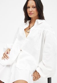 Balta lina blūze ar pūkainām piedurknēm, pogu priekšpusi un smalkām krokām plecu daļā. Apvienota ar apaļu sudraba kulonu.