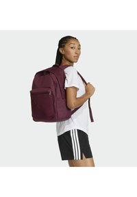 Zaino marrone con molteplici cerniere, spallacci imbottiti e una tasca frontale. Indossato sulla schiena di una persona con maglietta bianca e pantaloncini neri.