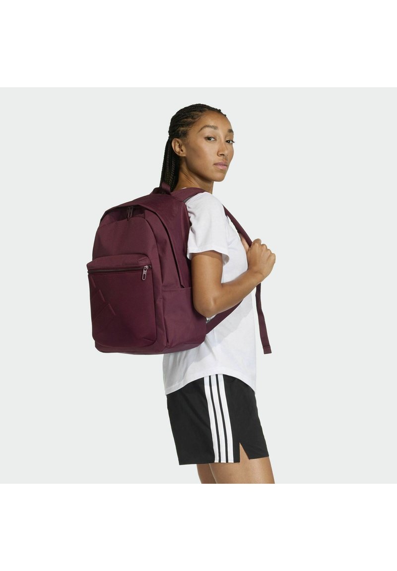 Zaino marrone con molteplici cerniere, spallacci imbottiti e una tasca frontale. Indossato sulla schiena di una persona con maglietta bianca e pantaloncini neri.