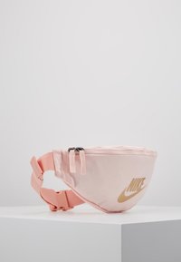 Růžový fanny pack Nike vyrobený z odolné tkaniny, se zakřiveným tvarem, dvěma zipov�ými přihrádkami a výrazným zlatým logem na přední straně.