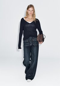 Frau geht und trägt einen schwarzen Strickpullover, ein weißes Unterhemd, weite dunkle Jeans, weiße Schuhe und hält eine braune Wildlederhandtasche.