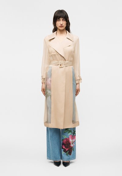 Femme portant un trench-coat beige avec des panneaux transparents sur un jean large orné de grandes impressions florales roses et rouges, debout devant un fond blanc.