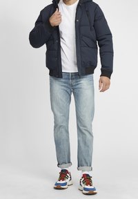 Granatowa puchowa kurtka z suwakiem z przodu, elastycznymi mankietami i kieszeniami, noszona na białej koszulce, zestawiona z jasnoniebieskimi dżinsami i wielokolorowymi trampkami.