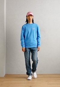 Lichtblauw sweatshirt met ronde hals, gecombineerd met donkere jeans en witte sneakers. Een roze pet maakt de outfit compleet. Eenvoudig, casual ontwerp.