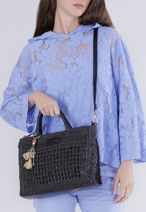 Nainen vaaleansinisessä kukallisessa pitsipaidassa, joka pitää kädessään mustaa punottua käsilaukkua, jossa on koristeelliset hapsut kahvoissa.