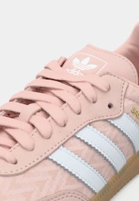 Ροζ σουέντ αθλητικό παπούτσι με ανοιχτό μπλε ρίγες, λαστιχένια σόλα, υφασμένο επάνω μέρος και λευκή ετικέτα με το λογότυπο της Adidas.